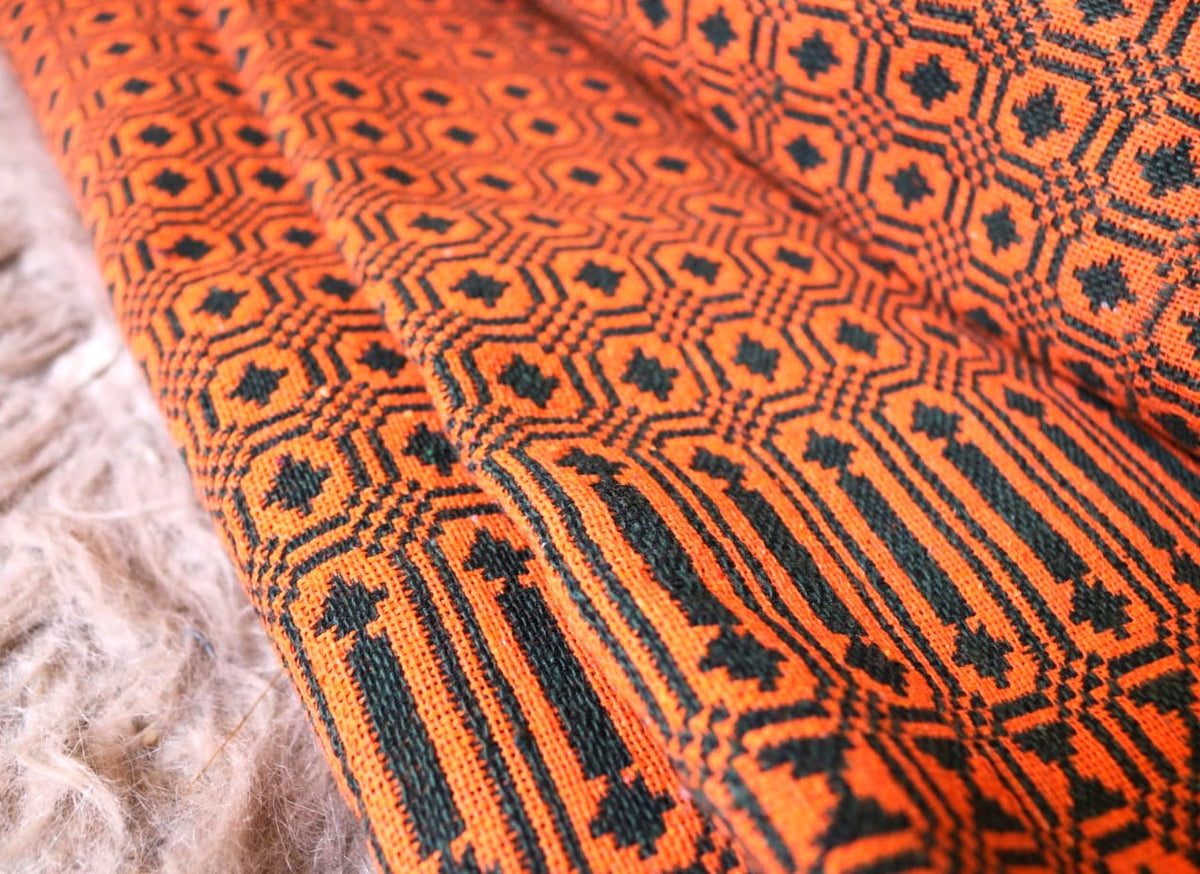 Close-up of a woven textile with a bold orange and black geometric pattern, shown draped over a soft beige textured surface.